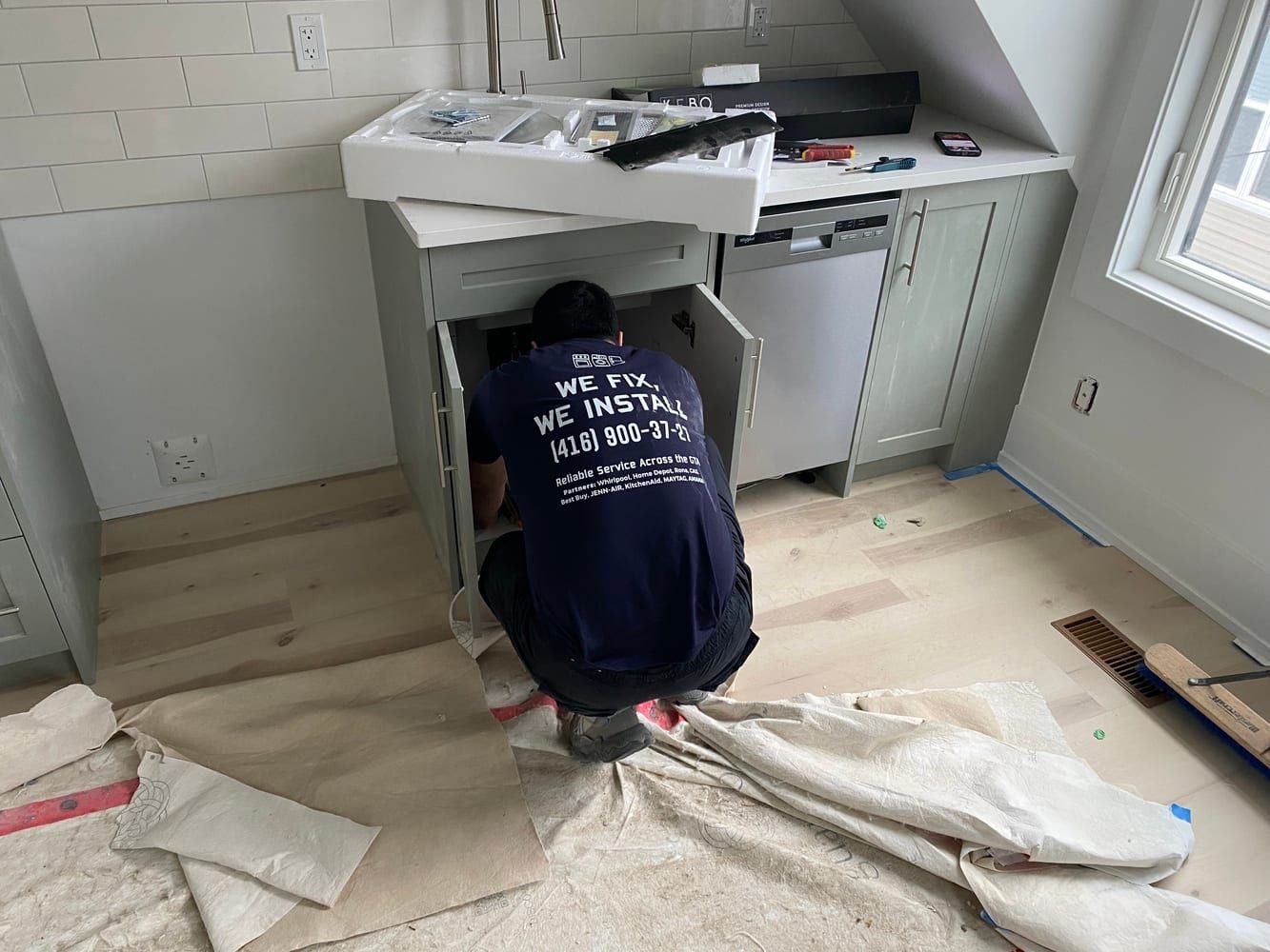 Appliance repair in Toronto — kitchen sink cabinet opened for a hookup, drop cloths over the new floor and tools laid out on the counter